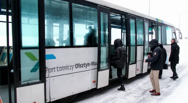 Szynobus z napisem "Port Lotniczy Olsztyn-Mazury" na tle śniegu, z pasażerami wsiadającymi.