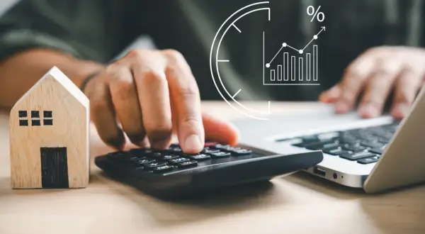 Person berechnet Miete unter Mietspiegel für das Finanzamt. Hausmodell, Rechner und Laptop im Fokus.