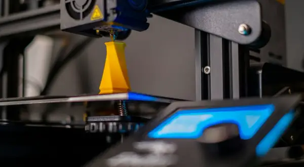 Drukarka 3D drukuje żółty obiekt, zbliżenie na dyszę i platformę roboczą.