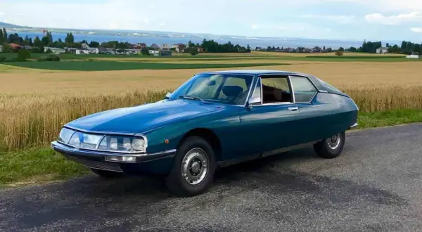 Piękny, turkusowy Citroen SM na tle złotego pola i jeziora. Klasyk motoryzacji w malowniczej scenerii.