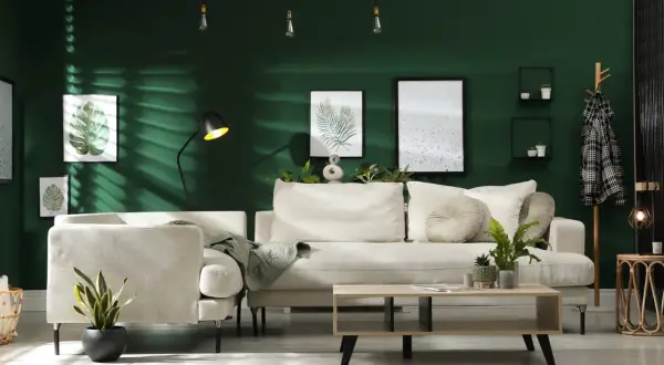 Ein gemütliches Wohnzimmer mit beiger Couch, Kaffeetisch und Pflanzen vor einer dunkelgrünen Wand. Ein Kleiderständer mit kariertem Mantel und ein trendiger Wandbehang vervollständigen das Bild.