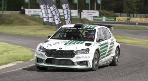 Biała Skoda Fabia Rally z zielonymi pasami pędzi po torze. Kierowca w kasku jest gotowy do akcji.