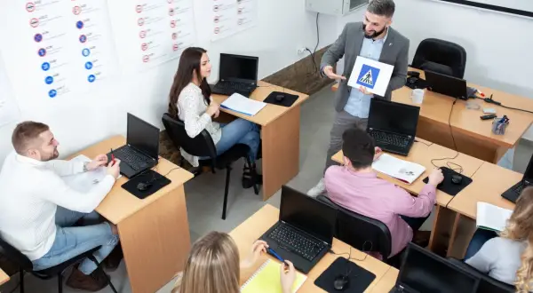 Nauczyciel pokazuje znak przejścia dla pieszych, omawiając zasady ruchu drogowego i kasowanie punktów karnych. Uczniowie słuchają, pracując przy laptopach.