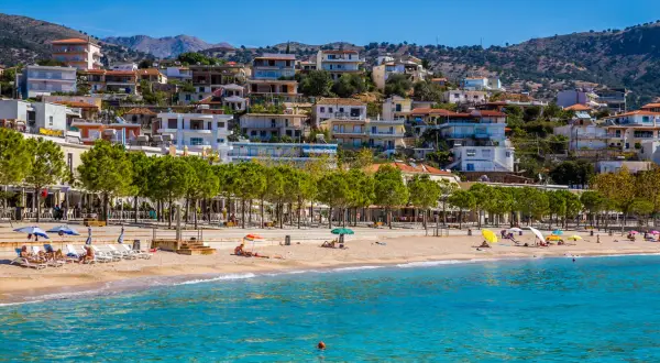 Piękna, słoneczna **Albania** w **czerwcu**. Ludzie odpoczywają na plaży, woda jest turkusowa, a miasteczko malowniczo wznosi się na wzgórzu.
