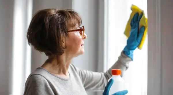 Mujer con gafas limpiando una ventana con un paño amarillo y guantes azules. La limpieza de exteriores es importante para mantener el hogar reluciente.