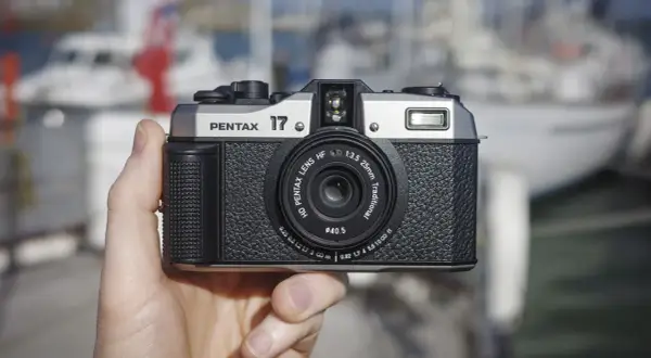 A hand holds the highest quality film camera, a silver and black Pentax 17, with a blurred marina in the background.