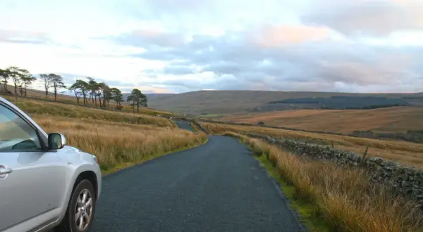 Voiture argentée sur une route sinueuse dans un paysage vallonné. La conduite à gauche est visible sur le côté de la route.