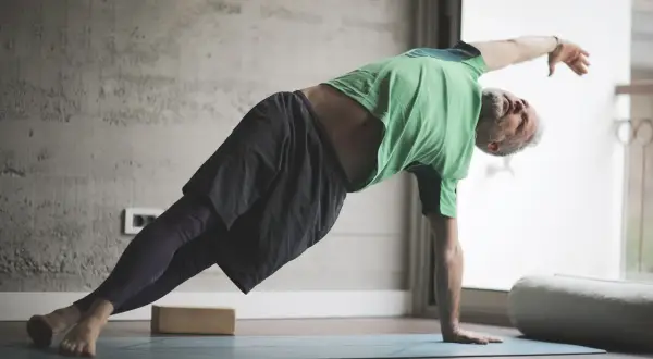 Hombre mayor con barba gris practica yoga para hombres en una esterilla, en postura de plancha lateral.