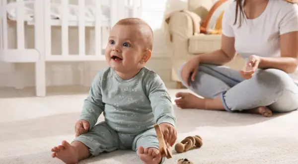 Uśmiechnięte 10 miesięczne dziecko bawi się drewnianymi zabawkami na dywanie, podczas gdy mama siedzi w tle.