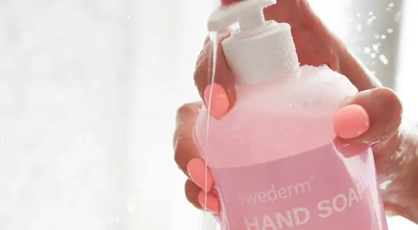 Dłoń z różowymi paznokciami trzyma różowy płyn do mycia rąk Swederm. Woda spływa po butelce, a na etykiecie widać napis "HAND SOAP". Czy gliceryna jest szkodliwa? Ten produkt zawiera jej wysoki poziom.