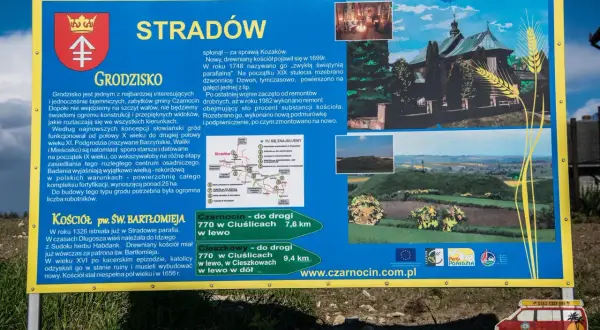 Tablica informacyjna o grodzisku Stradów, jego historii i kościele pw. św. Bartłomieja. Widok na krajobraz z wałami grodziska.