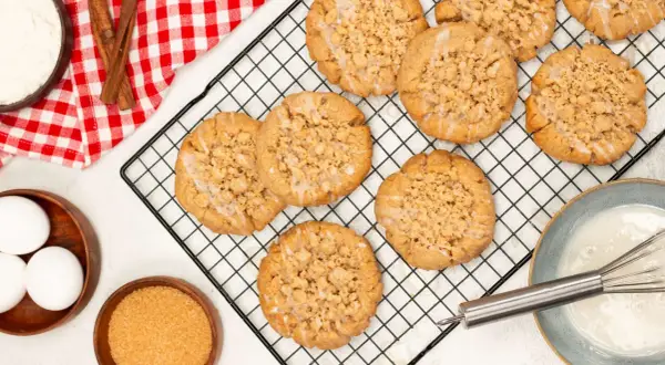 Frisch gebackene Zimt-Streusel-Kekse auf einem Kuchengitter. Zutaten wie Eier, Zucker und Mehl sind daneben zu sehen.