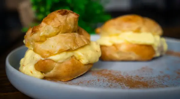 Zwei köstliche Windbeutel, gefüllt mit cremiger Vanillecreme, auf einem Teller mit Zimt bestäubt. Ein perfektes Windbeutel Rezept zum Nachmachen.