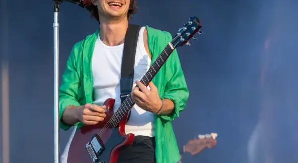 Joe Keery w zielonej koszuli i białej podkoszulce gra na czerwonej gitarze elektrycznej na koncercie.