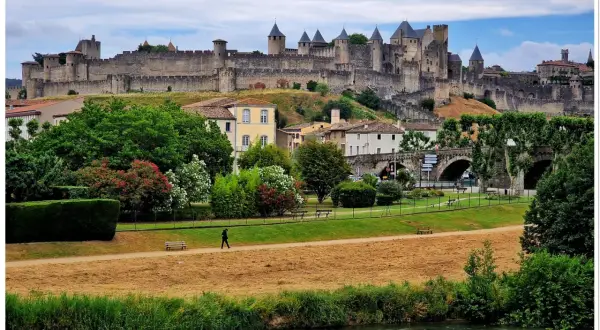 Majestatyczna twierdza Carcassonne góruje nad miastem. W dole zielony park, rzeka i spacerująca postać.