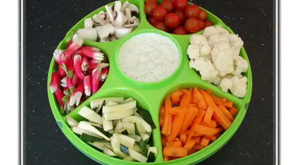 Plateau de crudités coloré : radis, champignons, tomates cerises, chou-fleur, courgettes et carottes, avec une sauce blanche au centre.