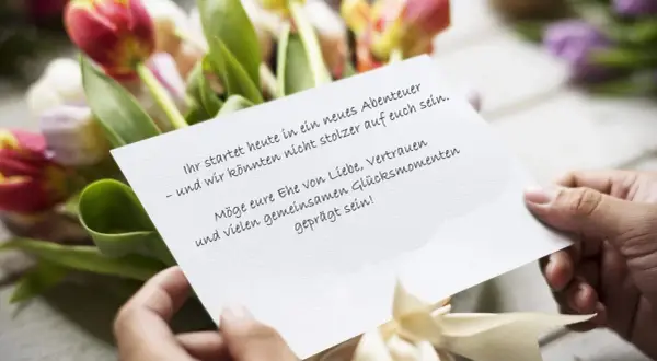 Hände halten eine Karte mit Hochzeitsglückwünschen vor Tulpen. "Ihr startet heute in ein neues Abenteuer – und wir könnten nicht stolzer auf euch sein. Möge eure Ehe von Liebe, Vertrauen und vielen gemeinsamen Glücksmomenten geprägt sein!