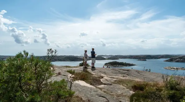 Två personer njuter av utsikten från Vetteberget i Fjällbacka. Vackert väder med moln och sol över skärgården.