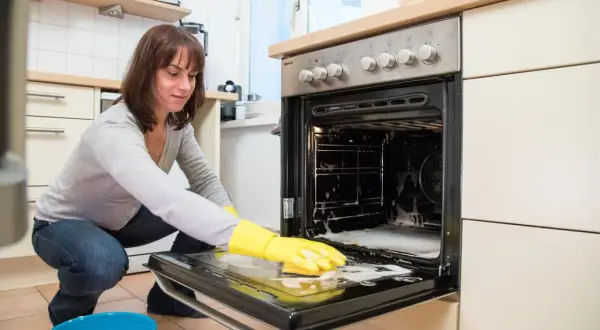 Frau mit gelben Handschuhen reinigt den Backofen. Schaum und ein blauer Eimer stehen bereit.