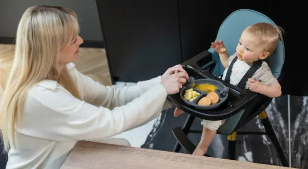 Mama karmi dziecko w krzesełku do karmienia, które nie siedzi samodzielnie. Na talerzyku jedzenie i ciasteczka.