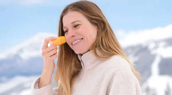 En hiver, faut-il mettre de la crème solaire ? Cette femme souriante en applique sur son visage, profitant d'un paysage enneigé.
