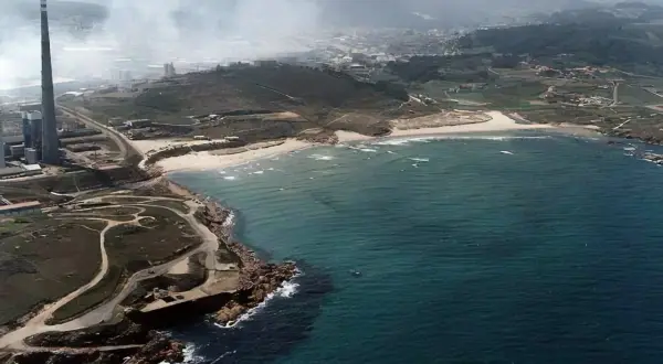 Vista aérea de la playa de Sabón, con una chimenea industrial y humo en el fondo, y el mar azul turquesa.