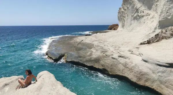 Una chica sentada en una roca en una cala chica, con el mar azul turquesa y acantilados blancos al fondo.