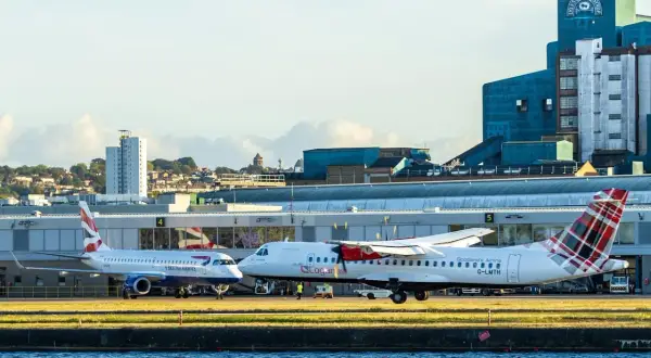 Zwei Flugzeuge, eines von British Airways, stehen auf dem Rollfeld. Im Hintergrund ist ein markantes blaues Gebäude mit der Aufschrift "Lyle's Golden Syrup" zu sehen. Welcher Flughafen London?