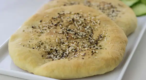 Två runda manakish zaatar med sesamfrön och örter på en vit bricka, med skivad gurka och tomat i bakgrunden.