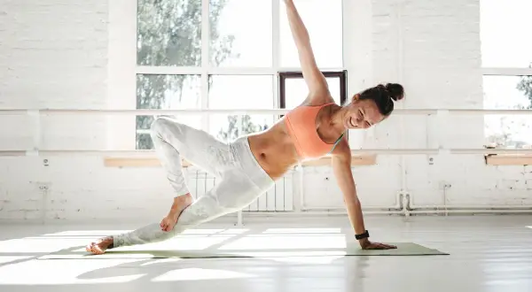 Donna sorridente in posa yoga dinamico, mostrando i benefici di forza e flessibilità in una luminosa sala da ballo.