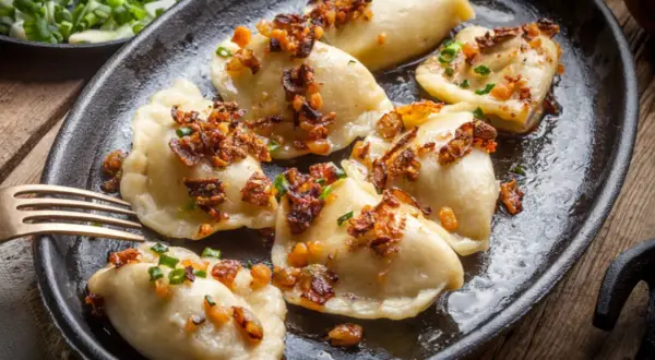 Pyszne pierogi z mięsem, posypane skwarkami i szczypiorkiem, podane na żeliwnej patelni.