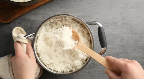 Hände halten einen Topf mit frisch gekochtem Reis. Eine hölzerne Kelle hebt eine Portion Reis heraus, perfekt auf welcher Stufe Reis kochen.