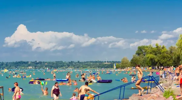 Tłum ludzi cieszy się słońcem i wodą na zatłoczonej balaton plaży. Wiele osób pływa, relaksuje się na materacach i bawi się w wodzie.