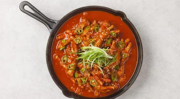 Kurczak w sosie gochujang, ozdobiony zieloną cebulką i sezamem, podany w żeliwnej patelni.
