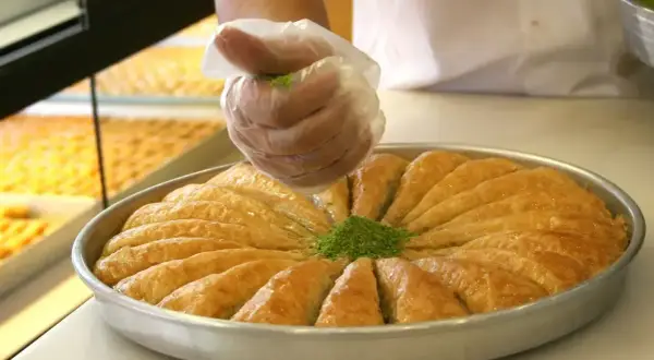 Ręka w rękawiczce posypuje pistacjami okrągłą blachę z baklavą. Tak wygląda baklava co to za przysmak!