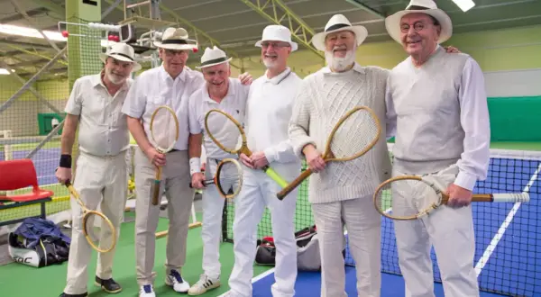 Sex äldre herrar i hattar och vita kläder poserar med tennisracketar, redo för en match i stil med Gustaf V:s tennis.