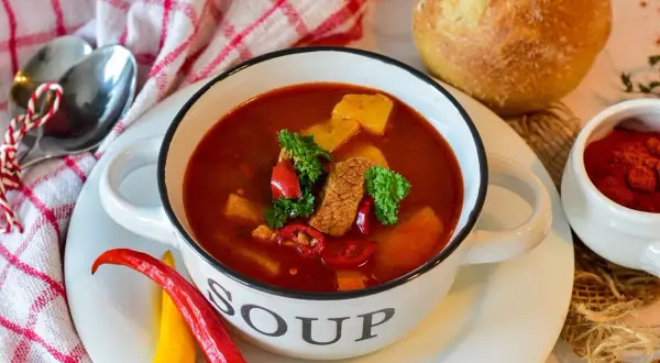 Miska gulaszowej zupy z mięsem, ziemniakami i papryczkami chili, obok chleb i przyprawy.