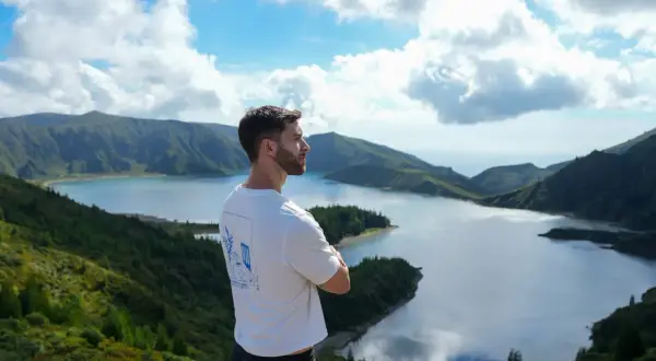 Hombre contemplando la **Lagoa do Fogo**, un lago azul rodeado de montañas verdes y un cielo nublado.