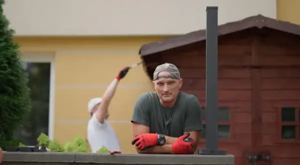Mężczyzna w czapce i rękawiczkach opiera się o mur. W tle inny mężczyzna maluje drewnianą szopę. Jak zgłosić się do polowania na ogród?