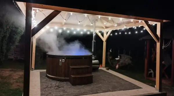 Drewniana beczka z gorącą wodą, z której unosi się para, pod zadaszeniem ozdobionym lampkami. Idealne miejsce na relaks w stylu sauna fińska.