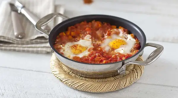 Un plat d'oeuf à la tomate mijoté dans une poêle, servi sur un dessous de plat en osier.
