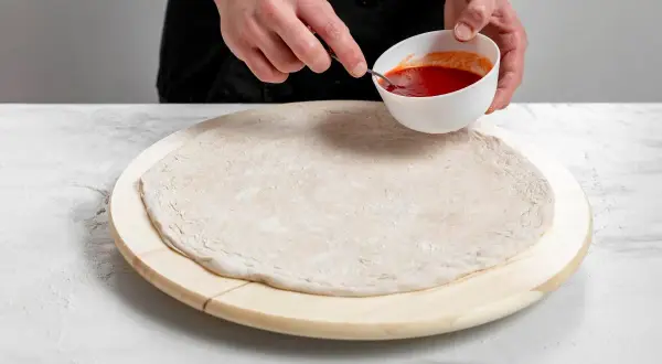 Ręce kucharza przygotowują szybką pizzę, nabierając sos łyżeczką z miseczki na surowe ciasto.