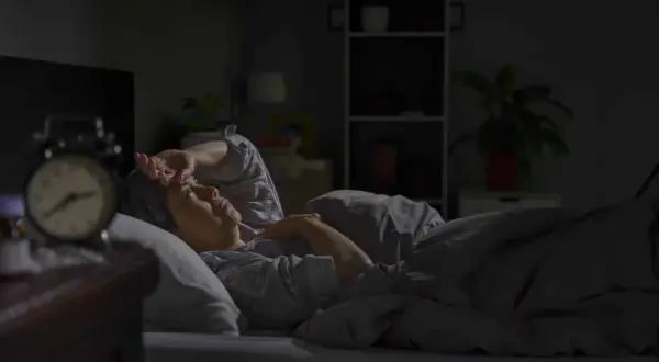 A person lies awake in bed, hand on forehead, experiencing insomnia after surgery. An alarm clock sits on the nightstand.