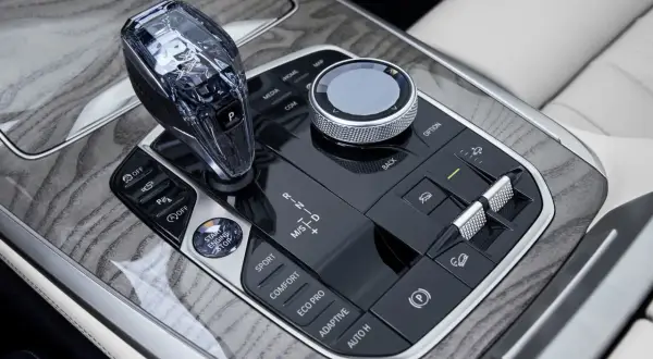 Kryształowa dźwignia zmiany biegów i pokrętło iDrive w BMW X7 2024, otoczone drewnianymi wstawkami i przyciskami trybów jazdy.
