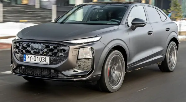 Nowe Audi Q3 w matowym, grafitowym kolorze sunie po drodze, prezentując swój sportowy charakter i nowoczesny design.