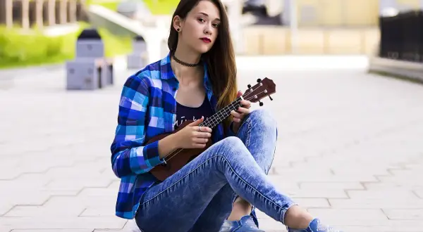 Dziewczyna w kratę i jeansach pokazuje, jak trzymać ukulele, siedząc na chodniku.