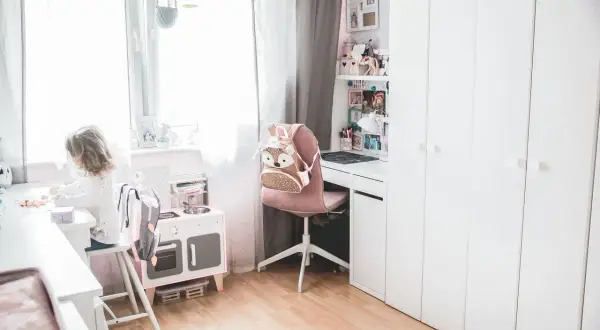Uroczy pokój dla 2 dzieci. Dziewczynka bawi się przy biurku, obok stoi różowa kuchnia i szafa.