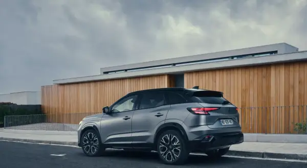 Vue arrière d'un SUV gris Renault Austral, stationné sur une route devant un bâtiment moderne en bois. La motorisation est performante.