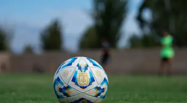 Piłka nożna z herbem Argentyny na trawie, w tle niewyraźne sylwetki graczy.