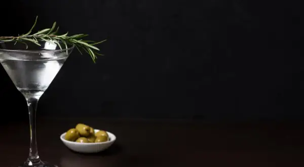 Elegancki drink z oliwką i gałązką rozmarynu, obok miseczka z zielonymi oliwkami.